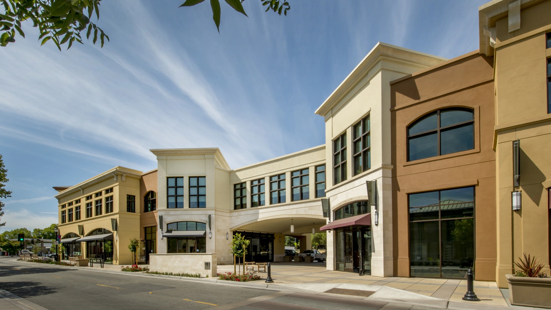 Street view of a modern, two-story shopping complex with large windows and an archway. Blue sky with wispy clouds above.
