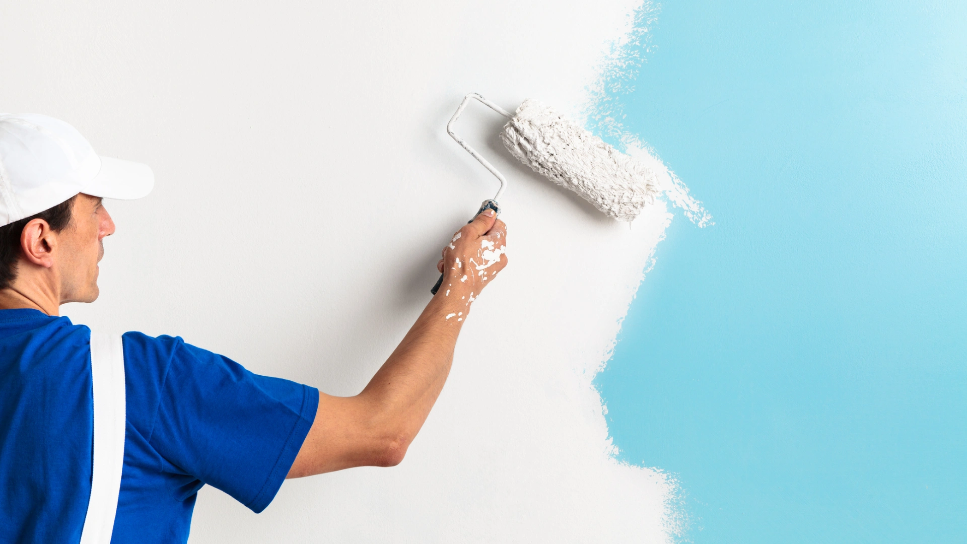 Person in blue shirt and white cap painting a white wall with a roller, covering a blue section.