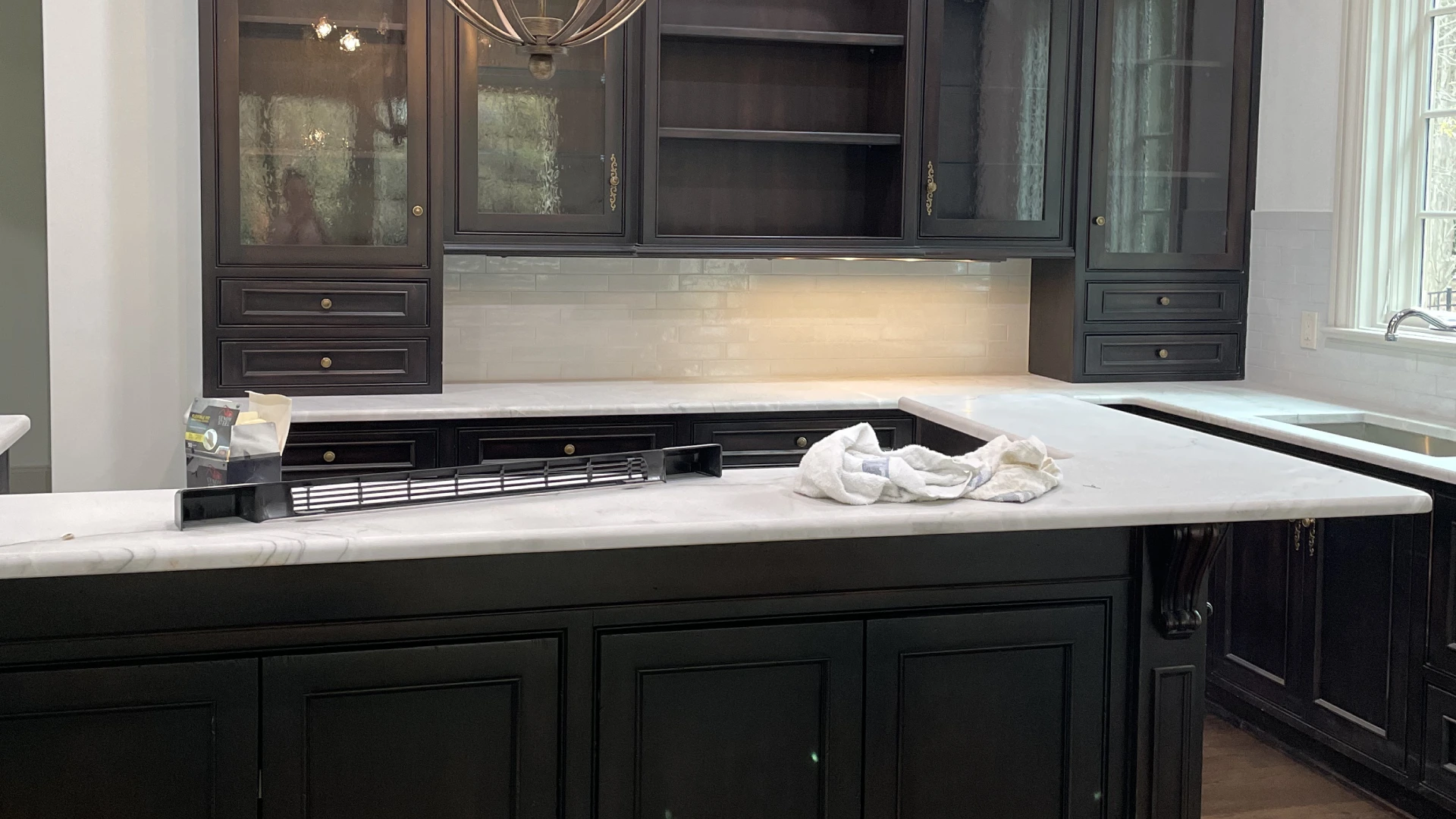 A kitchen with dark cabinets, a marble countertop, and an overhead light. A cloth and a tray rest on the counter near a sink.