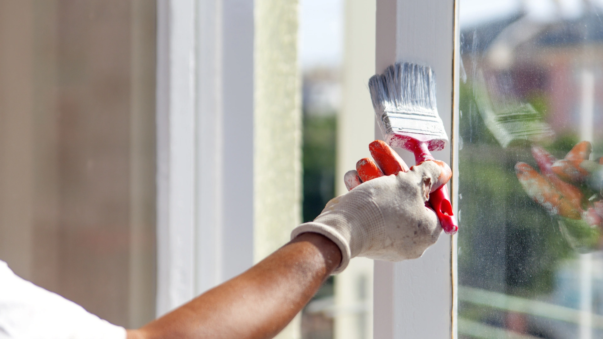 A person wearing a glove paints the edge of a window frame with a white paintbrush.