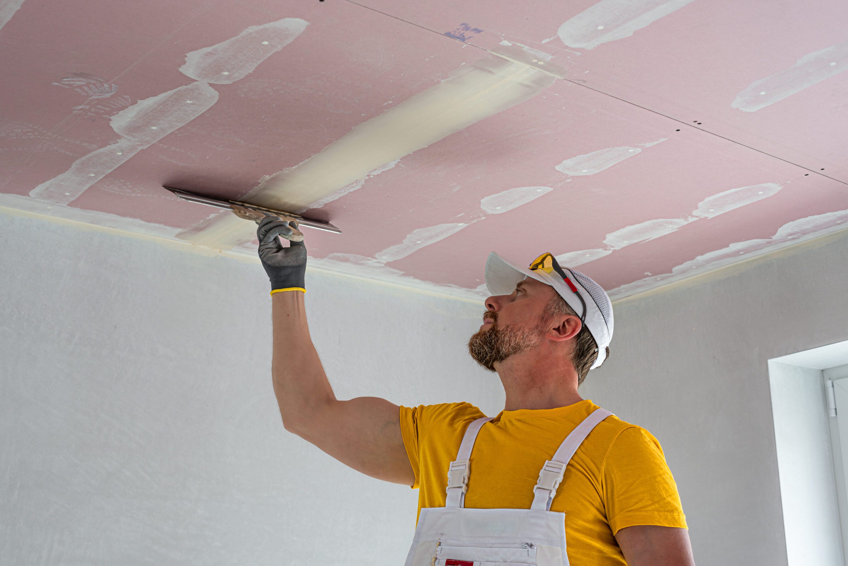 The worker make a plasterboard ceiling. He does taping plaster drywall ceiling joints. He is using special stainless steel taping knife.